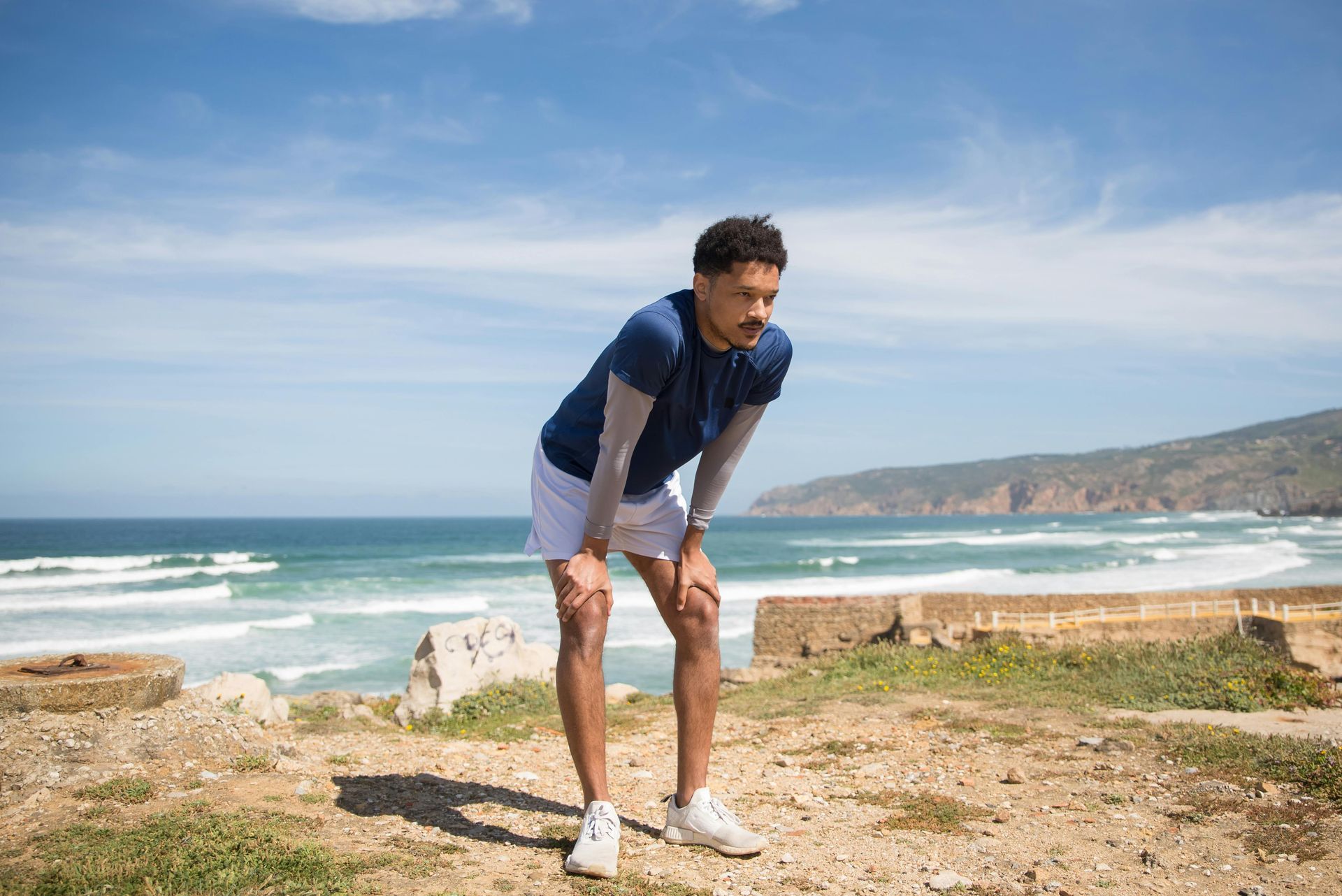 Man in athletic wear, hands on knees, appears winded near beach.