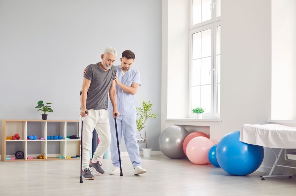 Man with cane, assisted by a healthcare worker, walks in a rehabilitation setting.