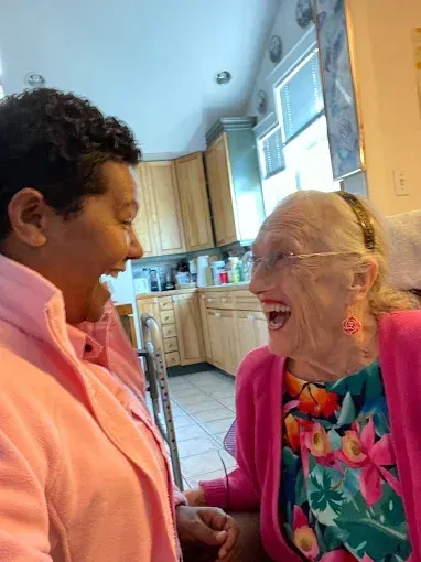Two people laughing, faces close. Woman in pink jacket, floral top; other in pink, indoors near kitchen.