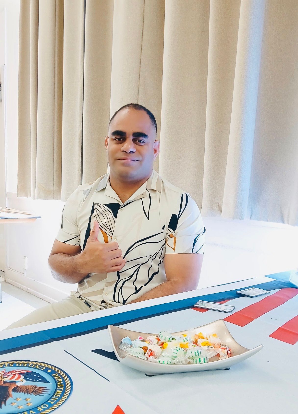 Man at table with food, giving thumbs up. Light skin, wearing printed shirt. Light background.