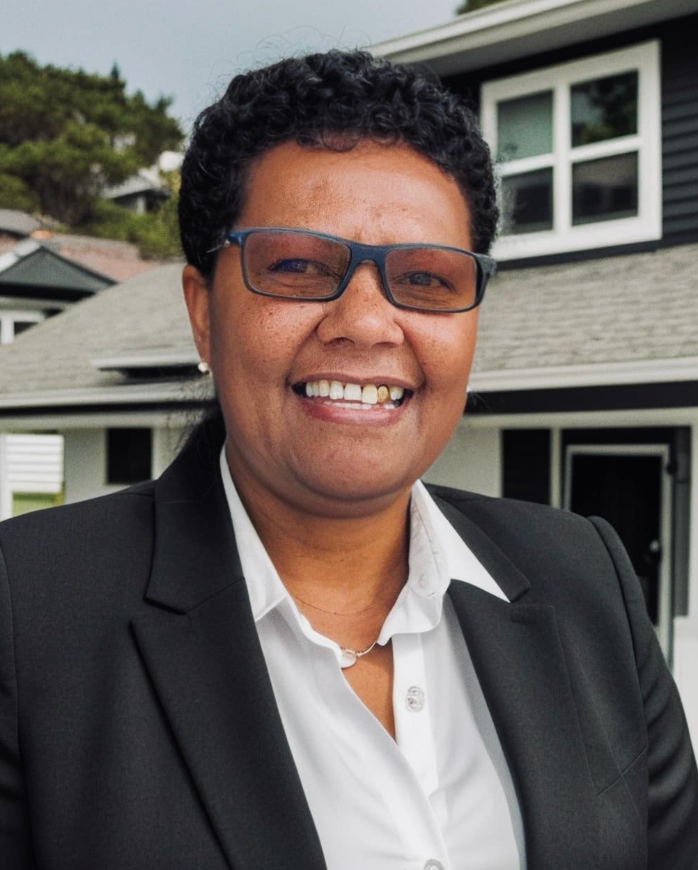 Woman in black suit smiles, wearing glasses, in front of a house.