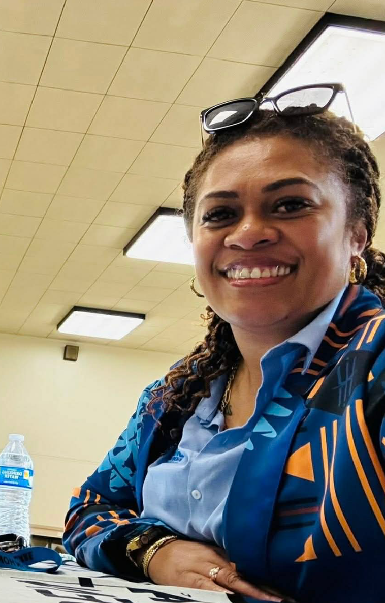 Woman smiles, wearing blue patterned jacket and glasses on head, sitting indoors near a water bottle.