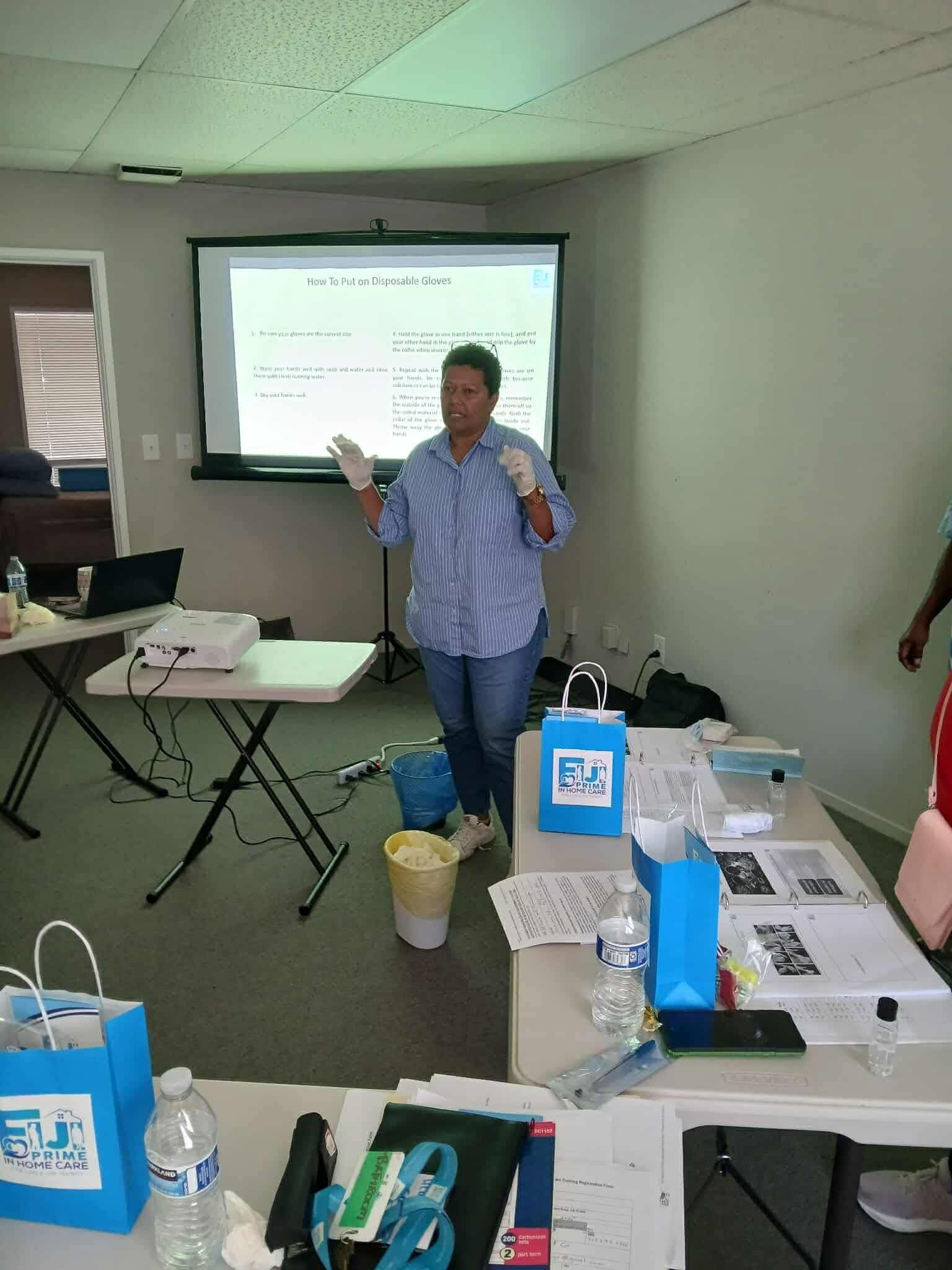 Person with gloves giving a presentation in a room. A projector displays a screen. Various items are on tables.