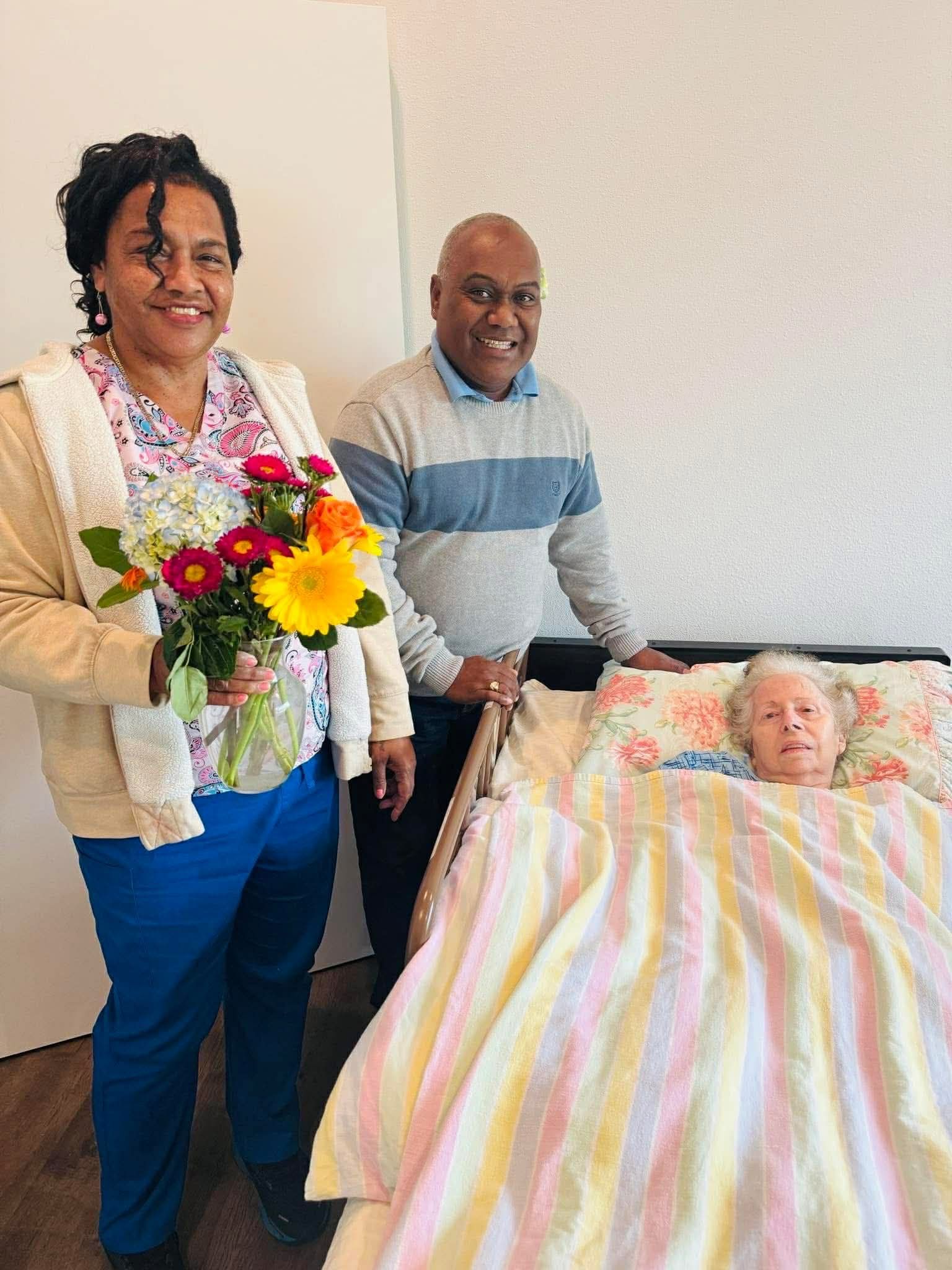 Woman holds flowers, smiles beside a man and a person in bed; soft colors, indoor setting.