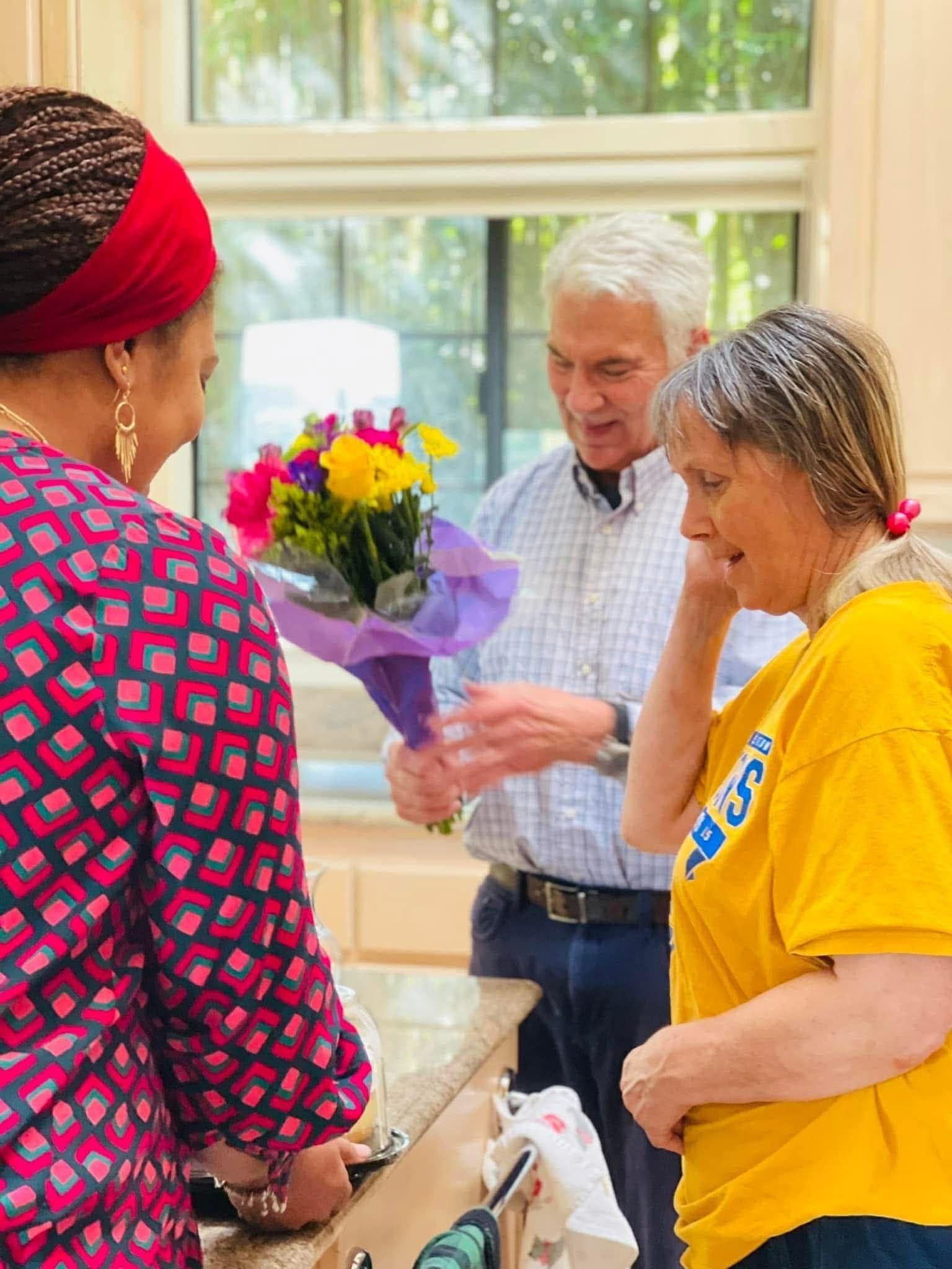 Man giving flowers to a woman; another woman watches. Kitchen setting with natural light.