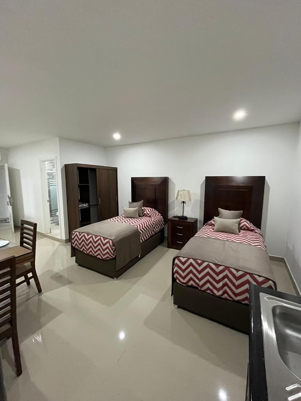 Dos camas individuales con cabeceros marrones en una habitación blanca, una cómoda y una mesa de comedor.
