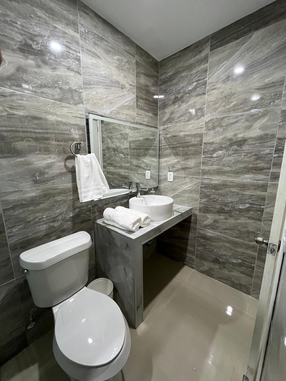 Baño moderno con paredes de azulejos grises, inodoro blanco, tocador de hormigón y lavabo redondo.