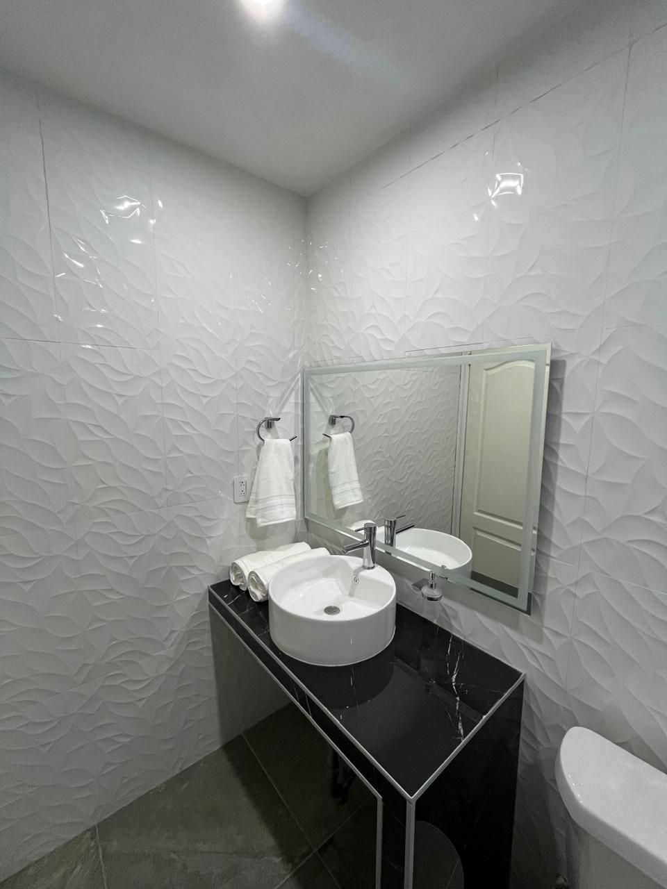 Pequeño baño blanco con paredes texturizadas, encimera negra, lavabo redondo, espejo, inodoro y toallero.
