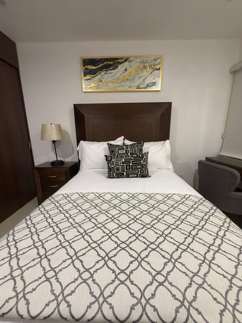 Dormitorio con cabecero marrón, mesita de noche y obras de arte. La cama tiene sábanas blancas, una manta estampada en gris 