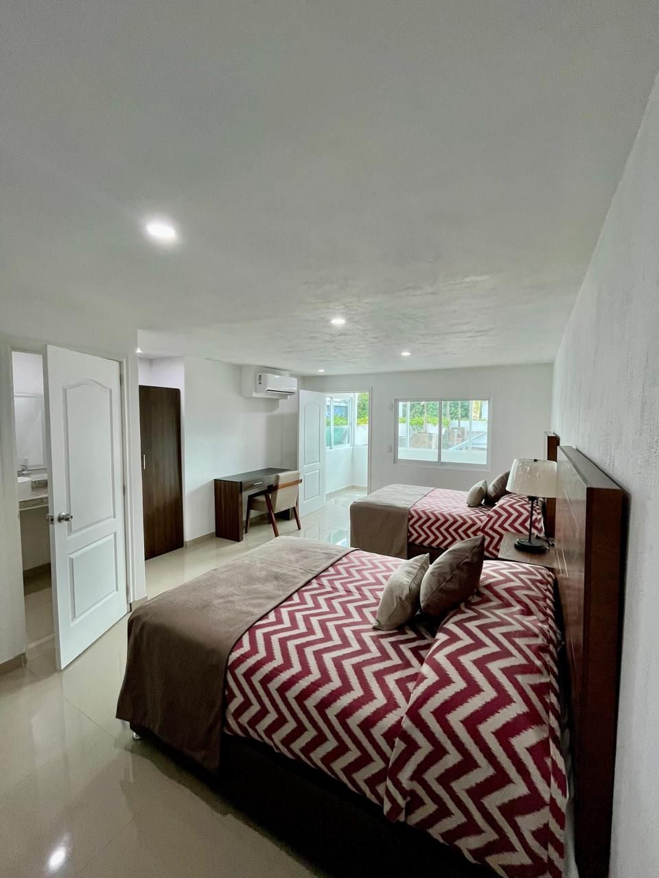Habitación de hotel con dos camas, escritorio y puerta abierta al baño. Habitación luminosa  ropa de cama roja en zigzag.
