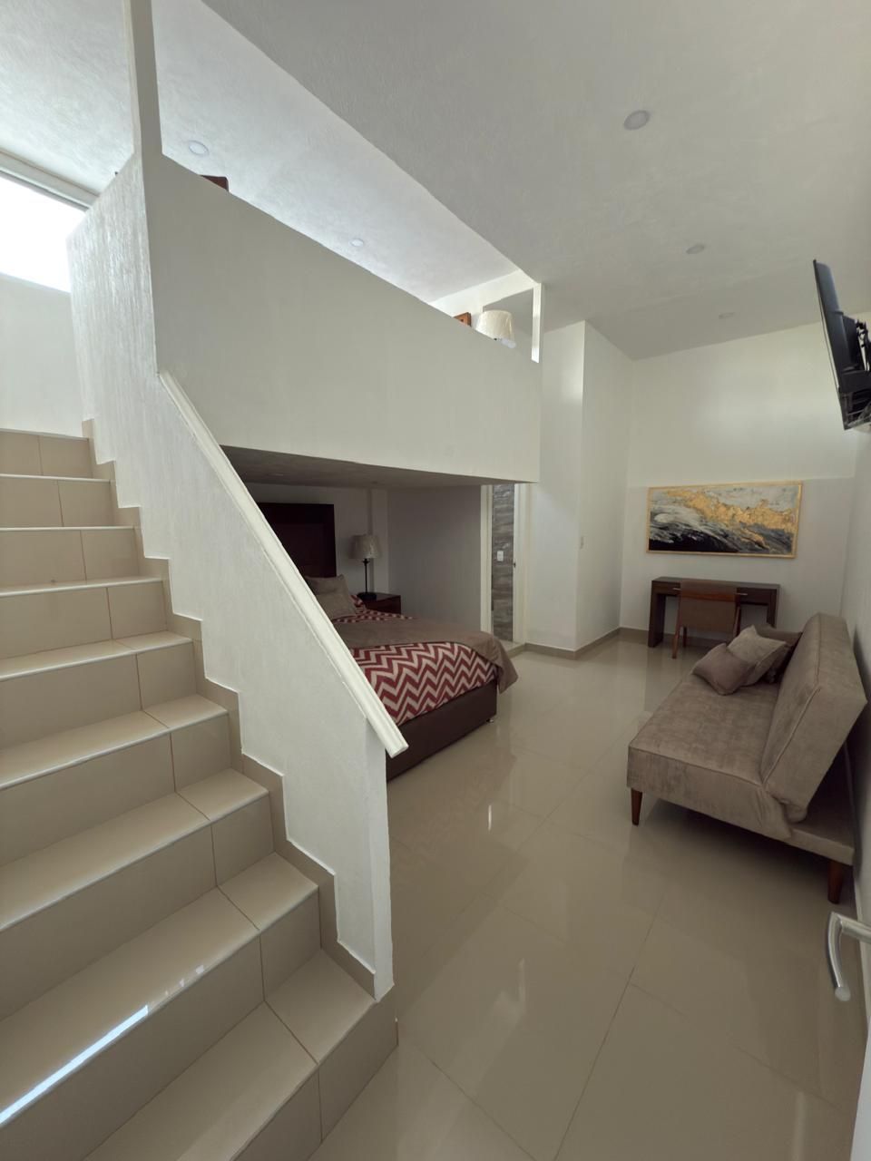 Dormitorio tipo loft blanco de dos plantas con escalera. Cama, sofá y obras de arte a la vista. Espacio luminoso y moderno.