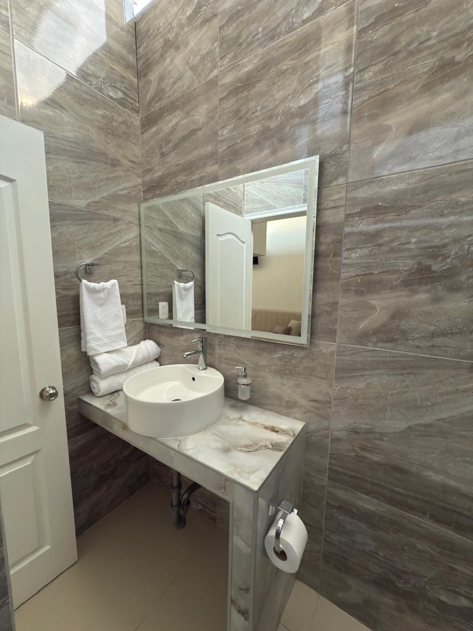 Baño con paredes efecto mármol, lavabo blanco, espejo y toallas, una puerta a la izquierda y un vistazo a otra habitación.