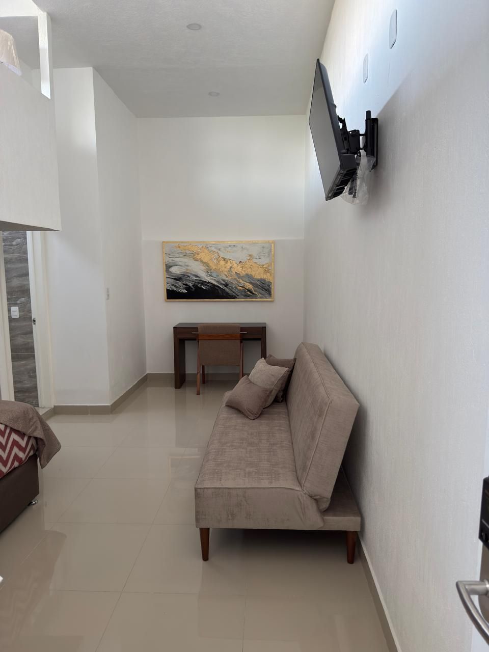 Pequeña habitación de hotel con sofá, escritorio y televisor montado en la pared contra una pared blanca.