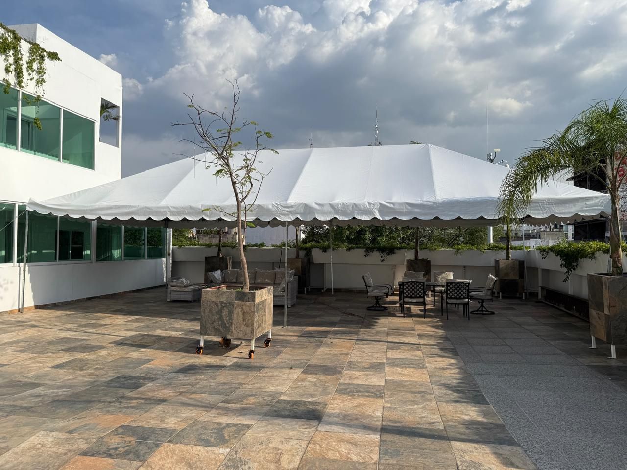 Carpa blanca sobre un patio al aire libre con asientos y vegetación cerca de un edificio con ventanas de vidrio.