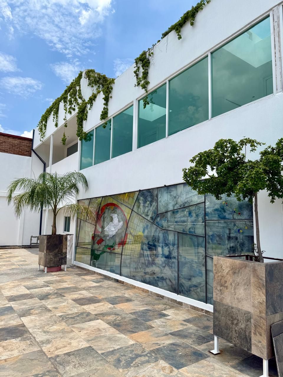 Edificio blanco con grandes ventanales, enredaderas y un panel de arte abstracto sobre un patio de ladrillo.