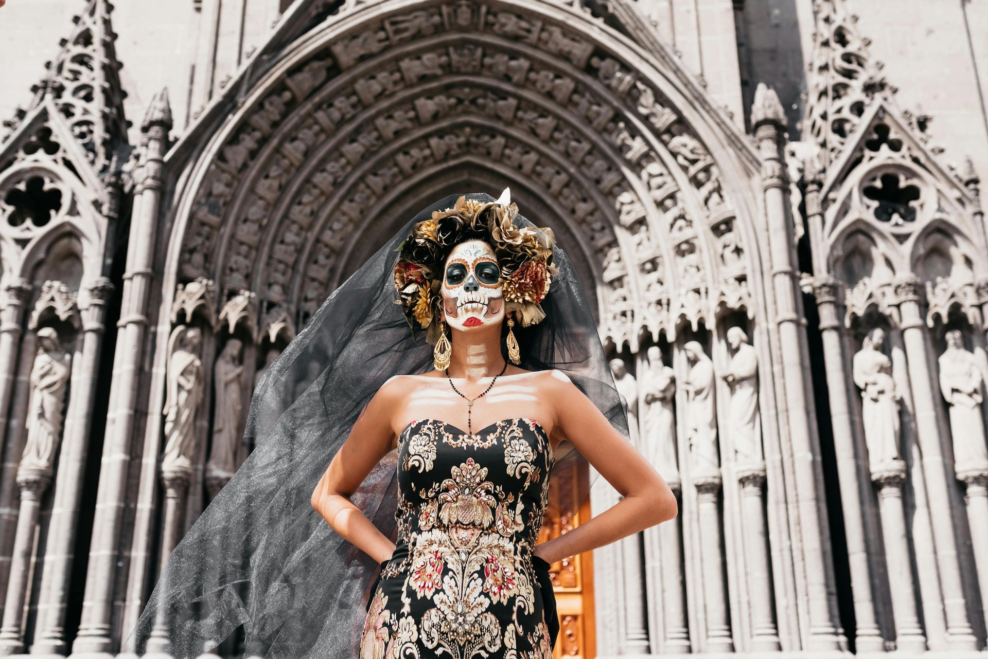 Mujer con maquillaje del Día de Muertos, corona floral, velo negro, vestido adornado, brazos en jarras, frente a catedral.
