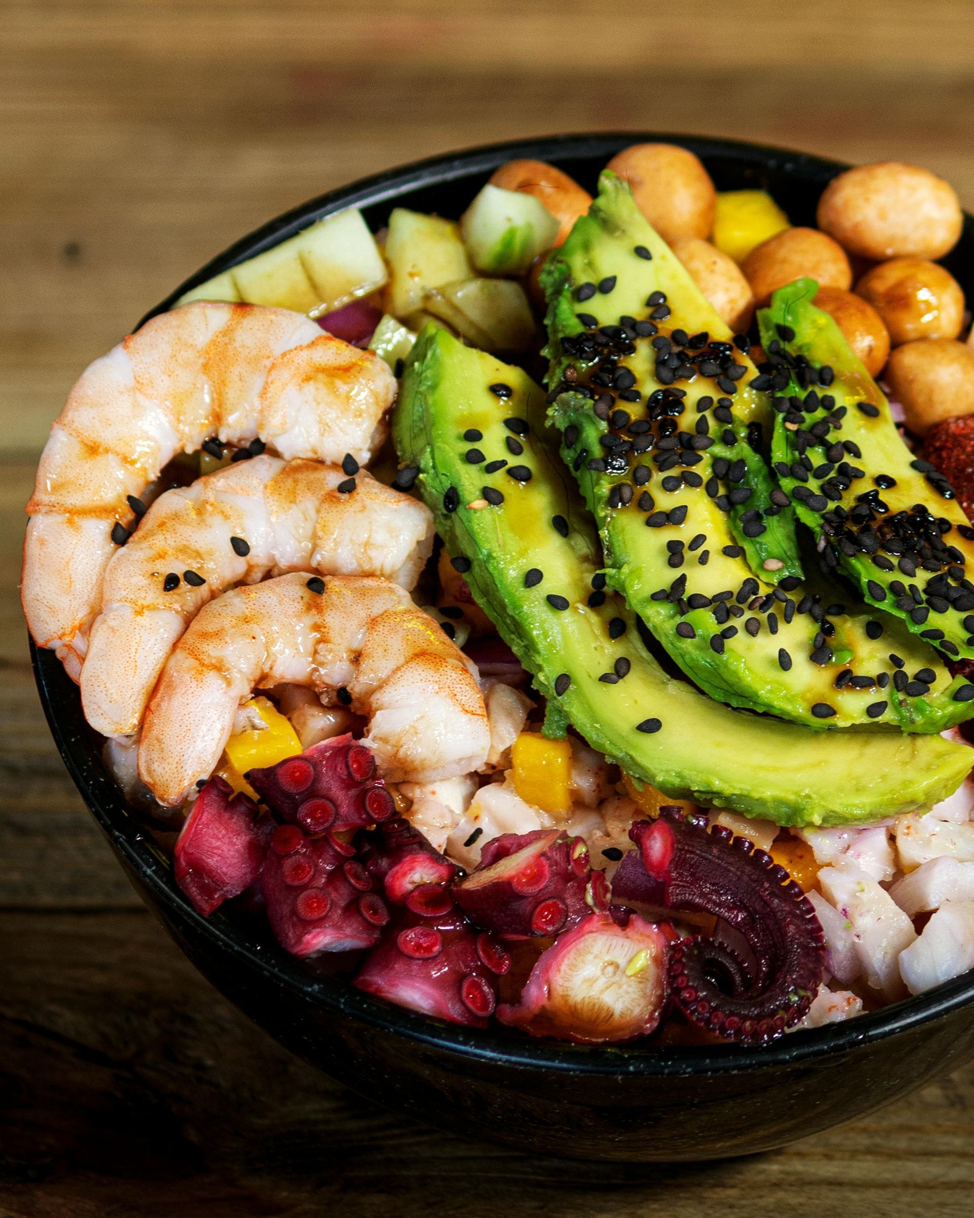 Poke bowl de camarones, pulpo y aguacate con semillas de sésamo negro y mango, en un recipiente negro.
