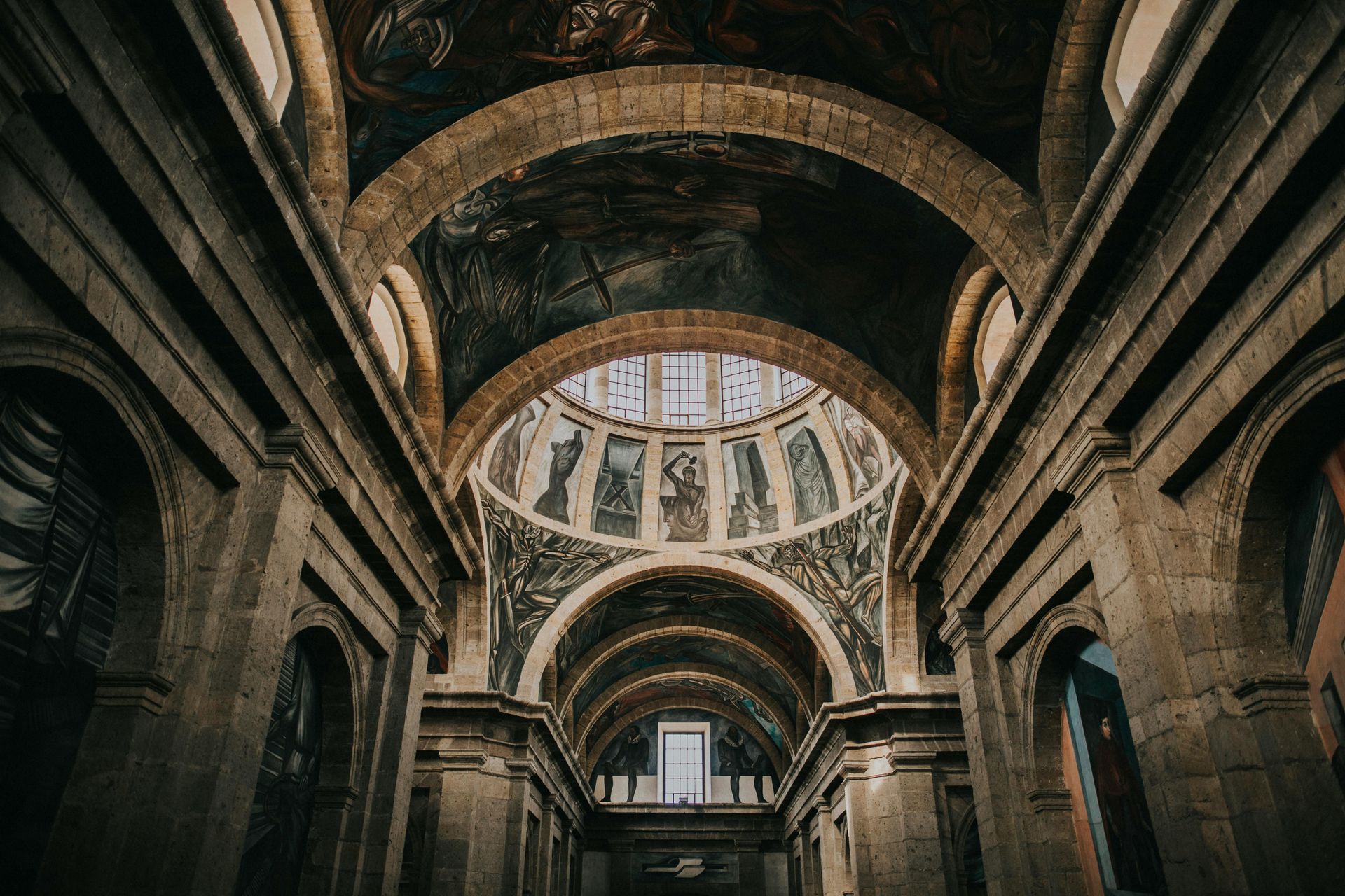 Arcos de piedra y techo abovedado en una gran iglesia ornamentada, con obras de arte pintadas en la cúpula.