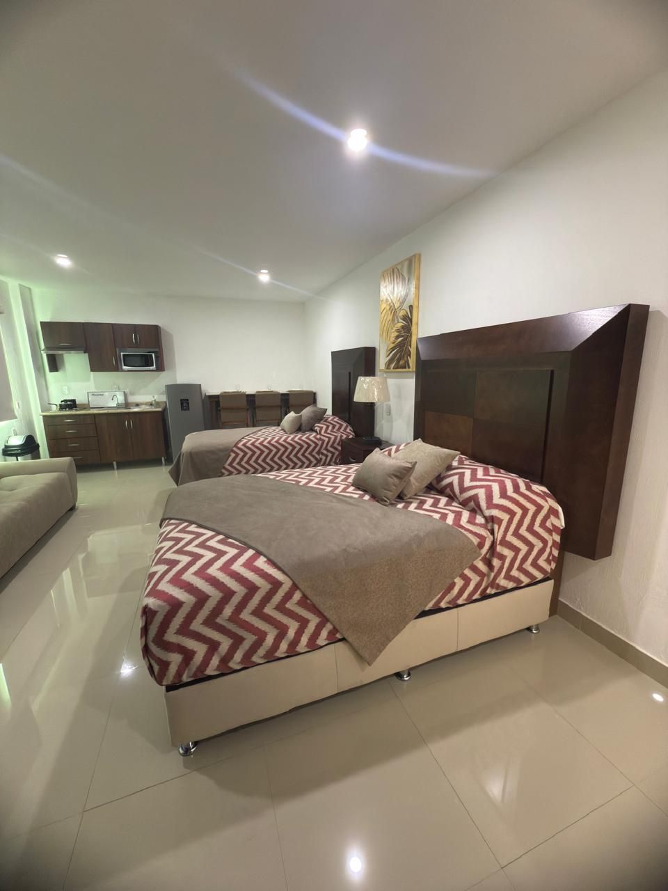 Una habitación de hotel bien iluminada con dos camas, una pequeña cocina y una zona de estar; tonos neutros y pisos pulidos.
