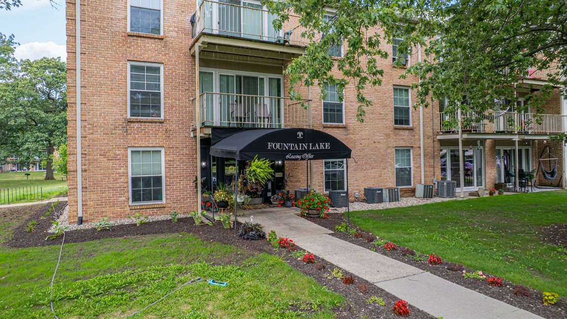 Brick apartment building with black awning and walkway. Green lawn with flowerbeds.
