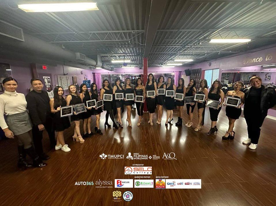 Group of women in black dresses holding signs, posing in a dance studio.