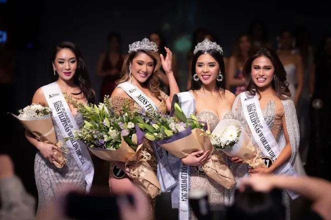 Four beauty queens in crowns and sashes holding bouquets, smiling, on a stage.