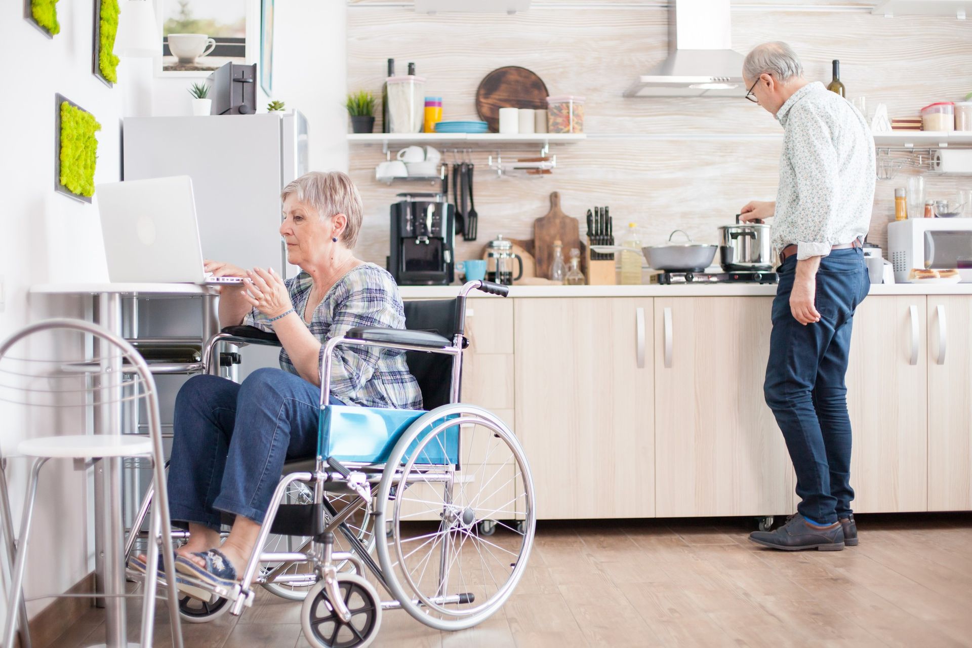 An elderly woman in a wheelchair is using a laptop while a man cooks in the kitchen.