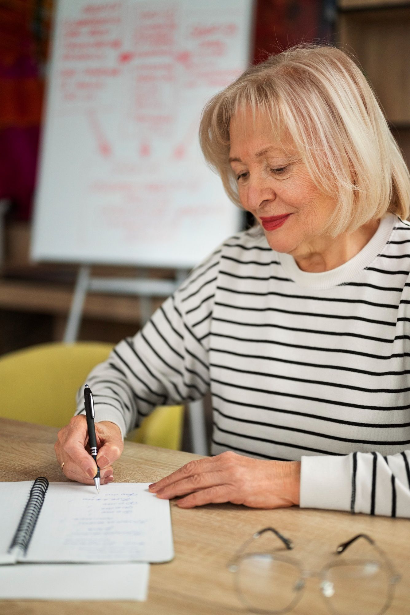 An elderly woman is sitting at a table writing in a notebook.