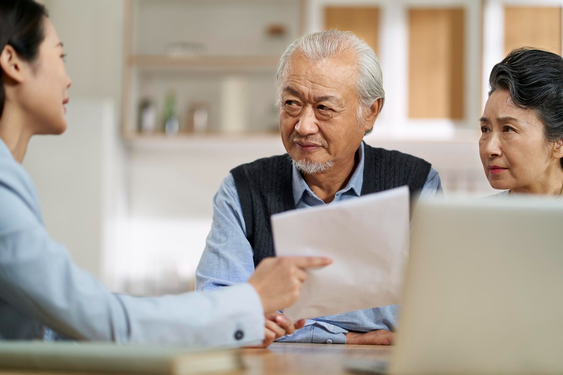 An elderly couple is sitting at a table with a woman pointing at a piece of paper.
