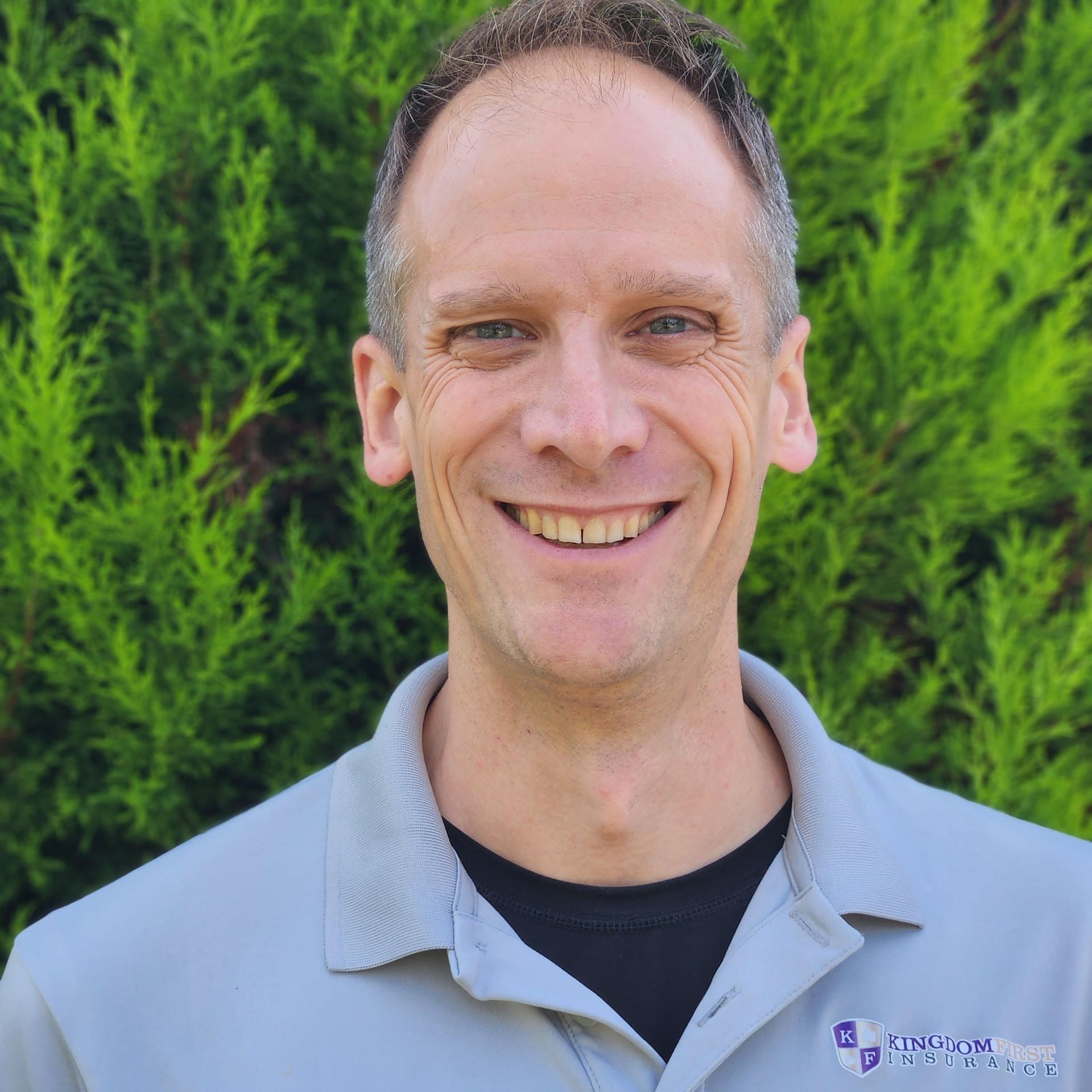 A man in a grey shirt is smiling in front of trees