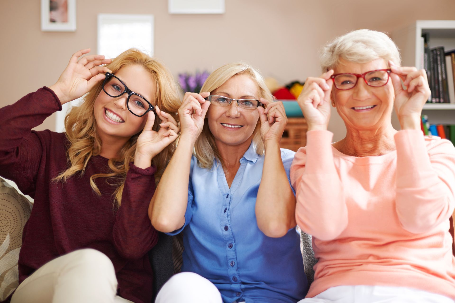 Three women are sitting on a couch trying on glasses.