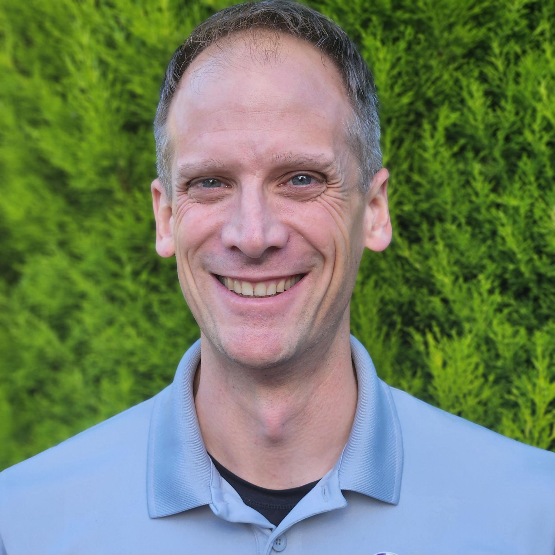 A man in a grey shirt is smiling in front of trees
