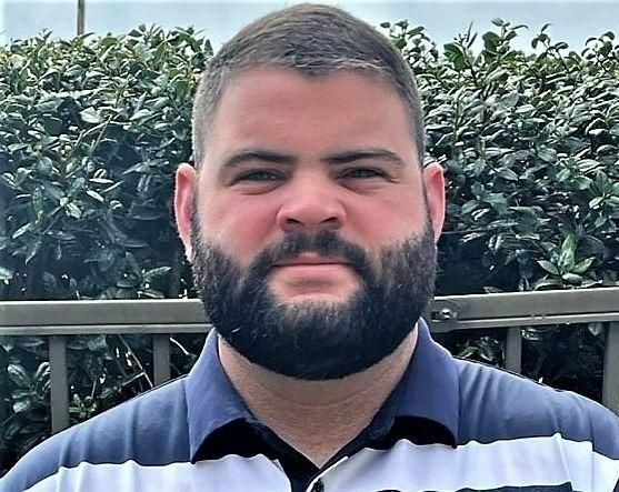 A man with a beard is standing in front of a fence.