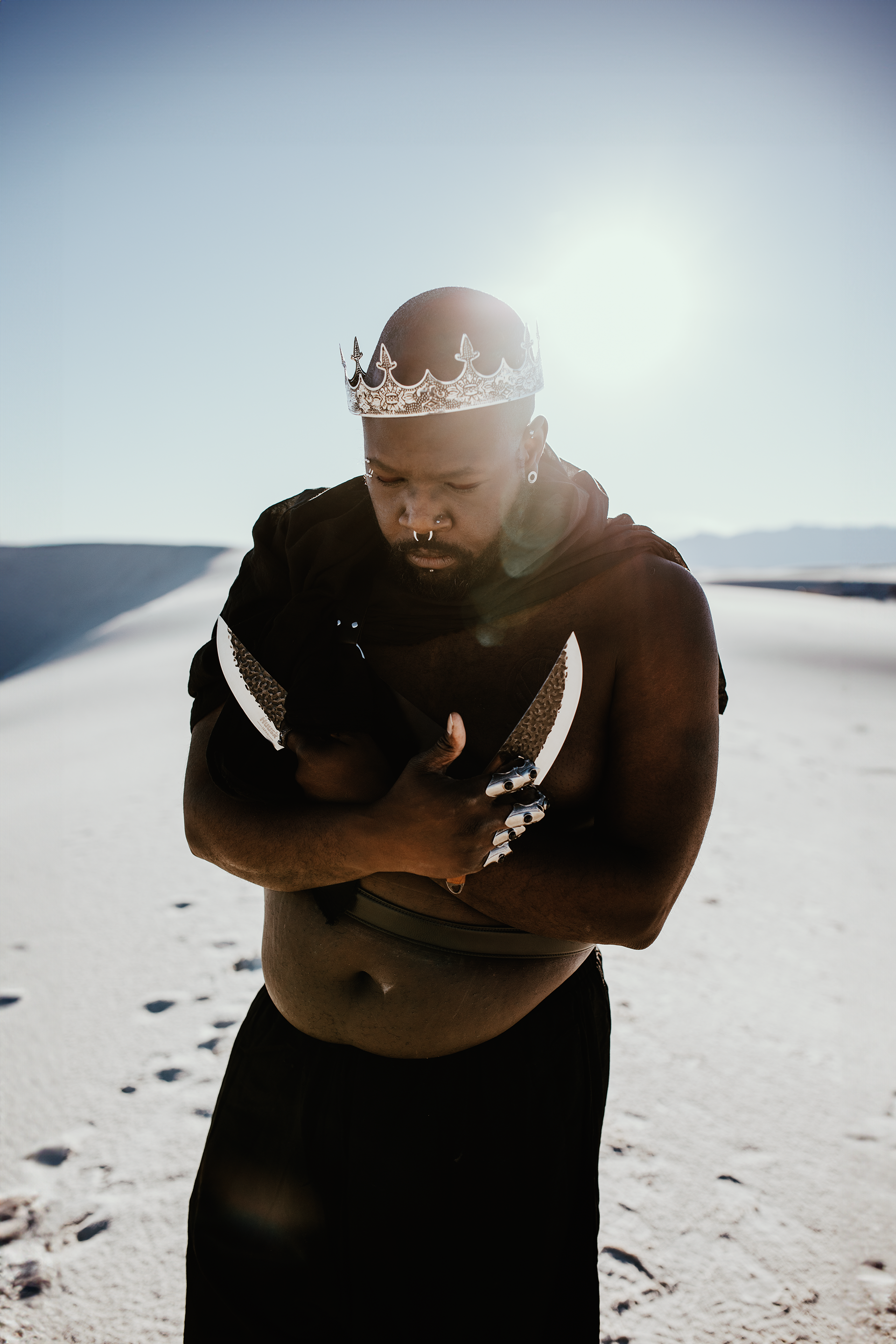 Black man wearing a crown, holding knives in a desert. Sunlight shines.