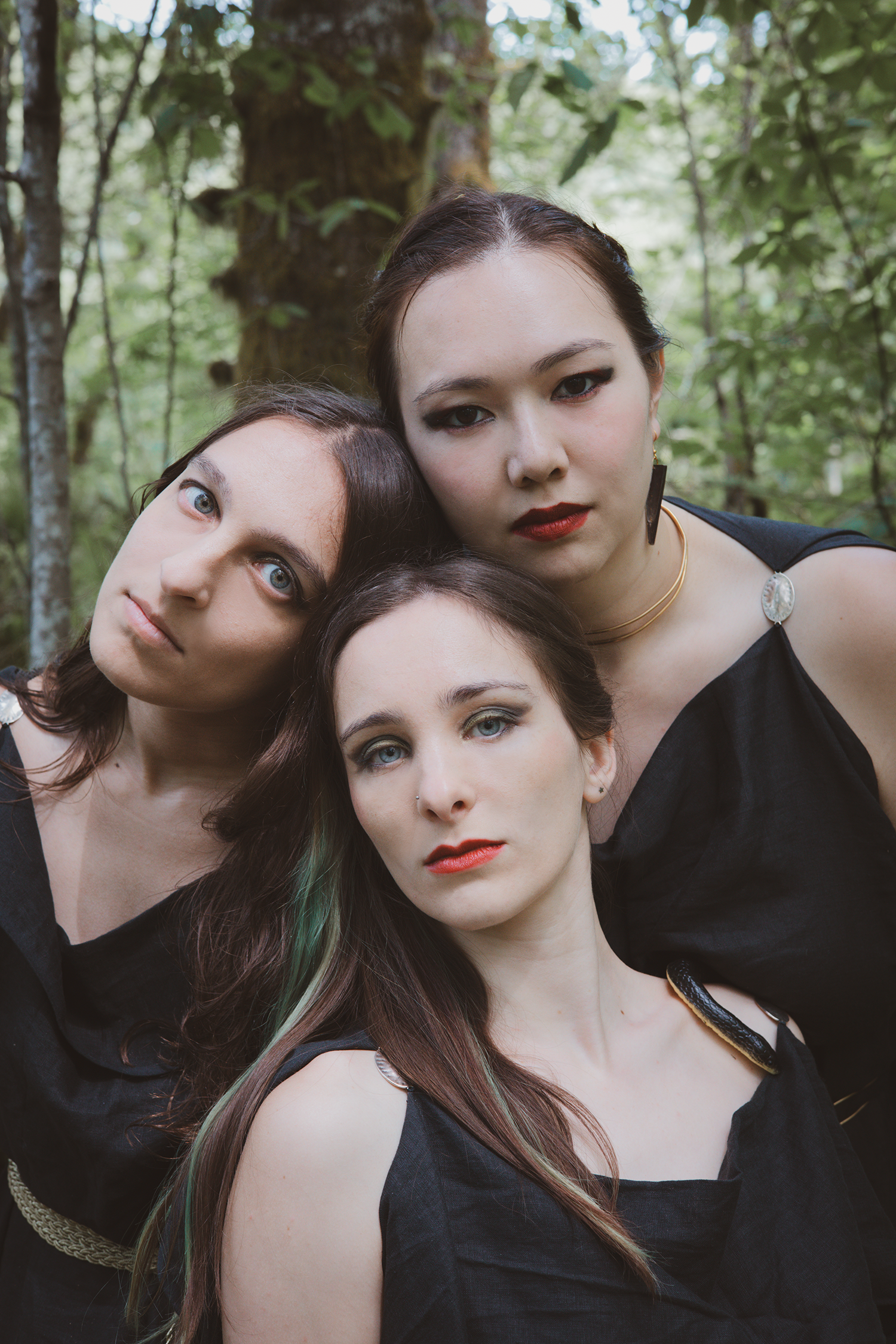 Three women in black dresses pose in a forest.