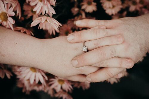 Hands clasped, showing engagement ring, pink flowers in background.