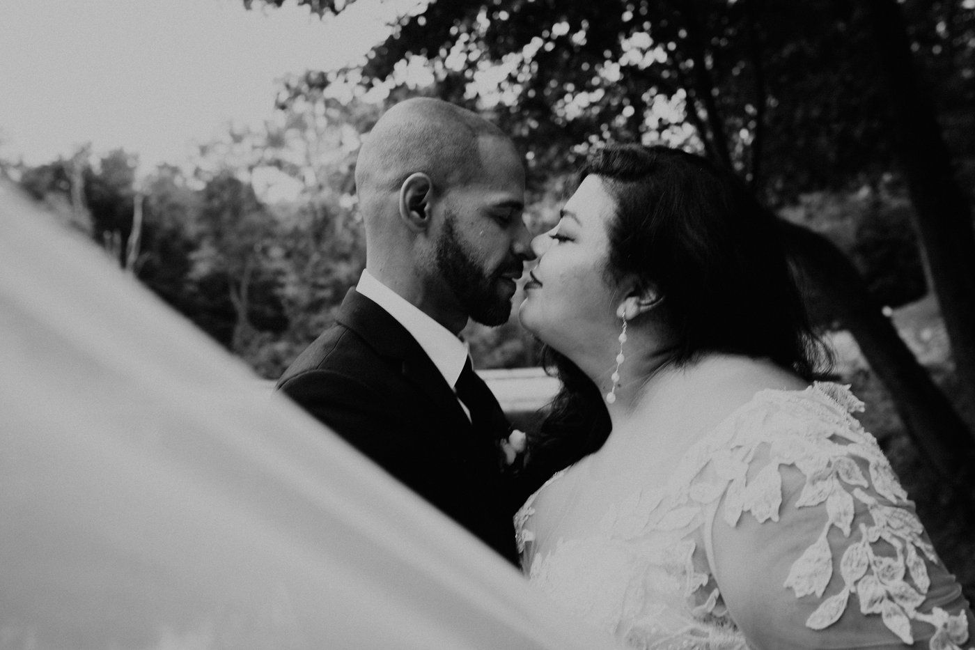 Couple about to kiss; bride in a lace dress, groom in a suit, veil blowing, trees in background.