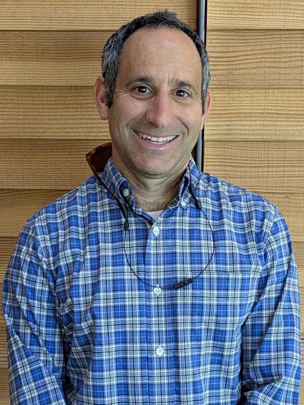 Man in blue plaid shirt smiles at the camera, glasses around his neck, standing in front of a wood-slat wall.