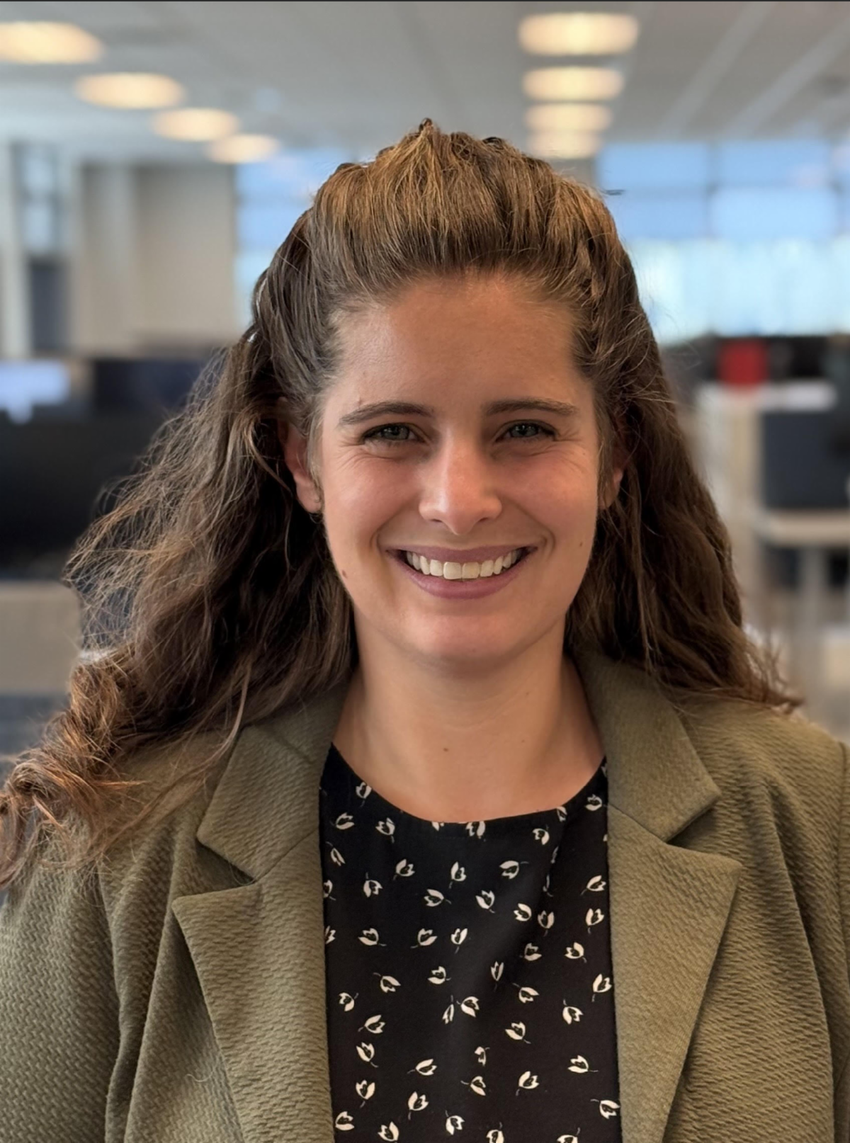 Woman with brown hair smiles, wearing a blazer over a patterned top. Indoors, blurred background.