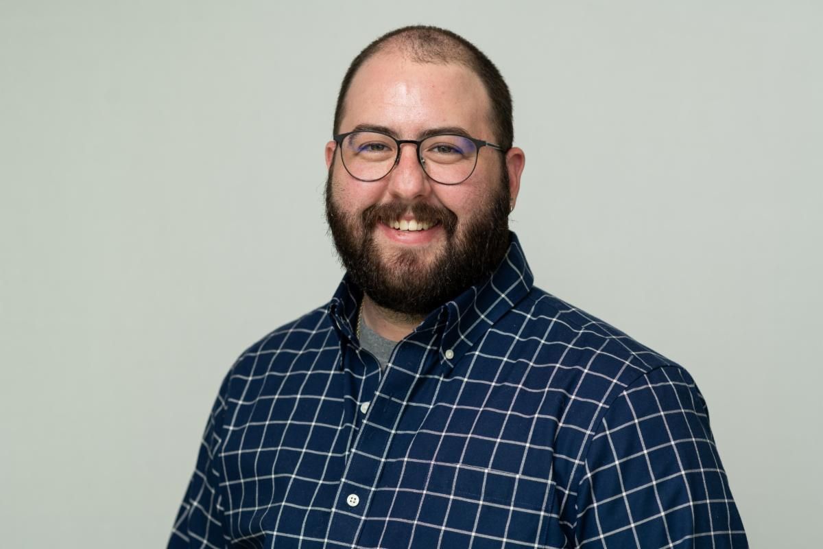 Man with a beard and glasses smiling, wearing a blue plaid shirt, against a white backdrop.