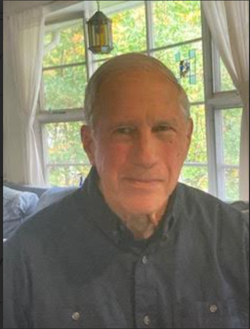 Man in a dark blue collared shirt smiling indoors, with a window and curtains in the background.