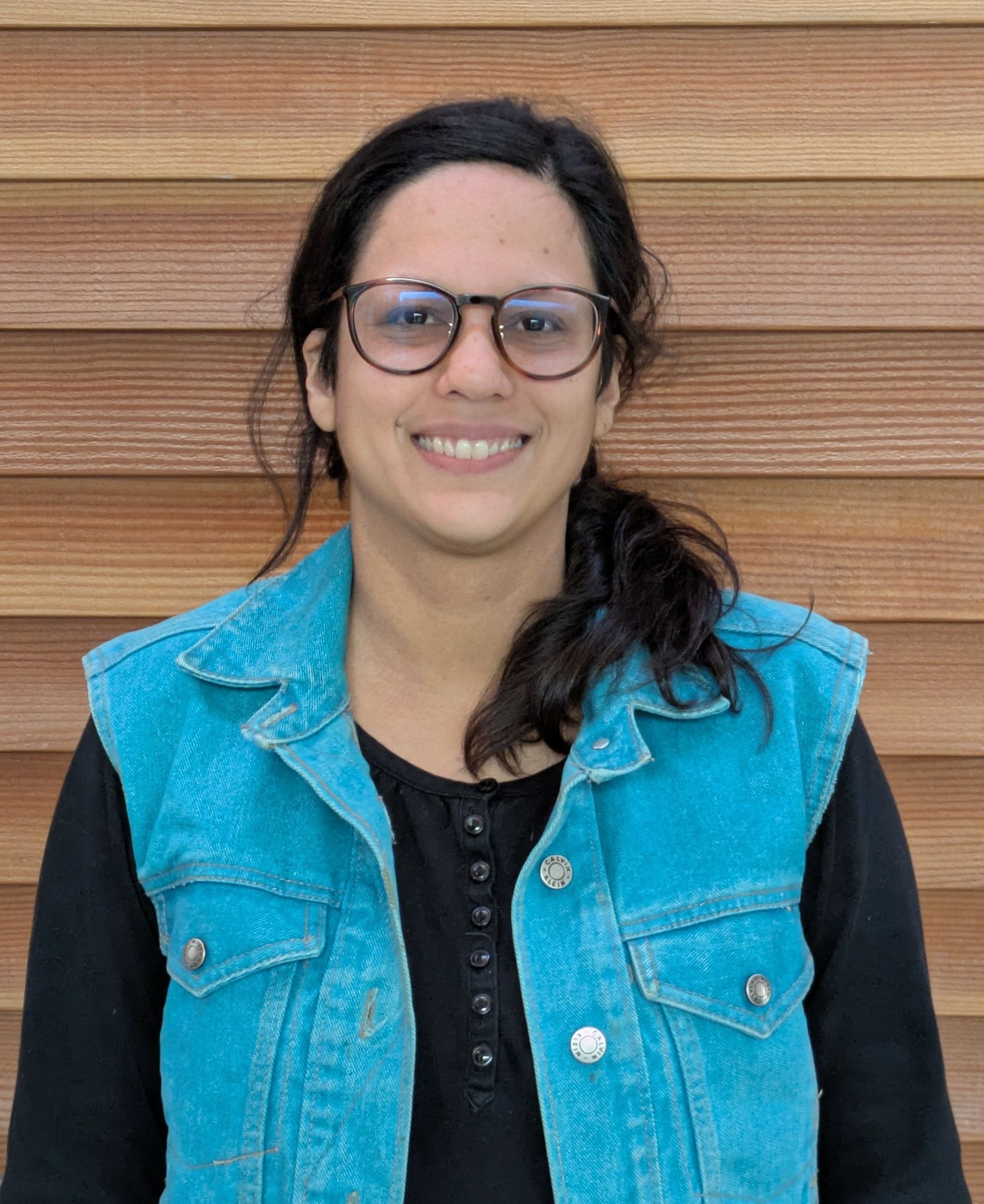 Woman in glasses and denim vest smiles in front of a wood-paneled wall.