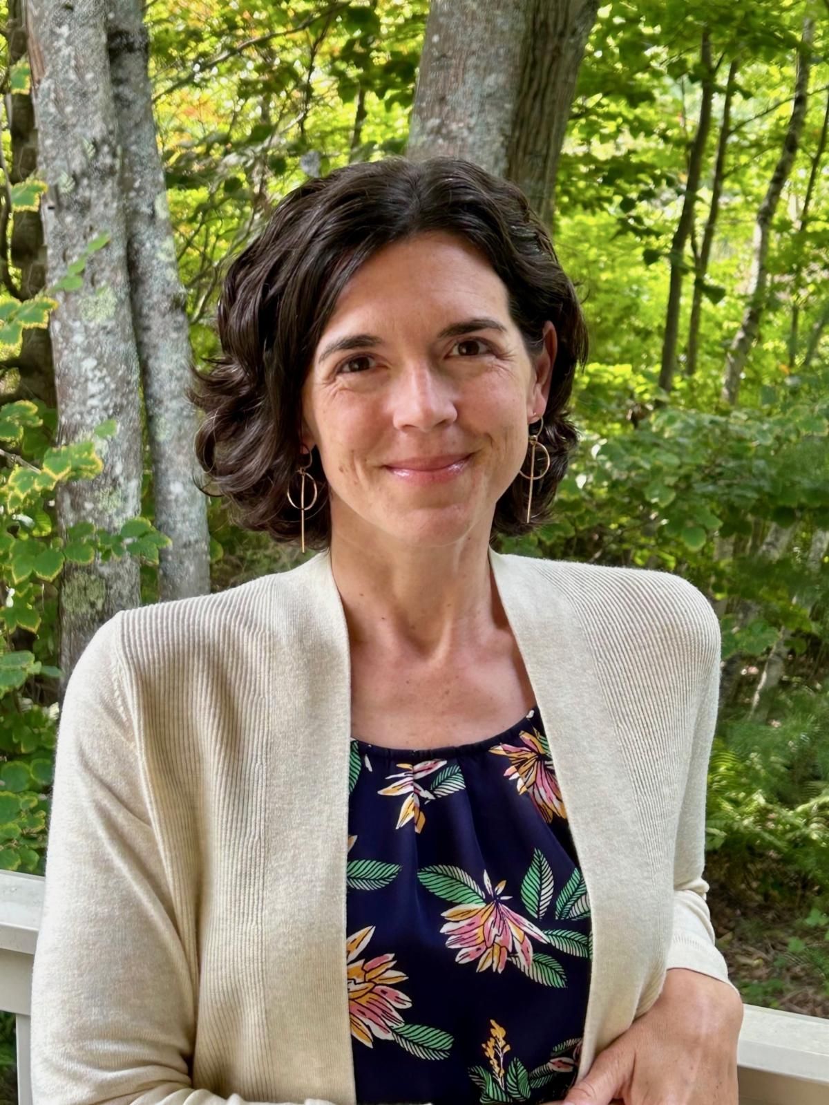 Woman with dark hair smiles, wearing a cream cardigan over a floral print top, standing outside.