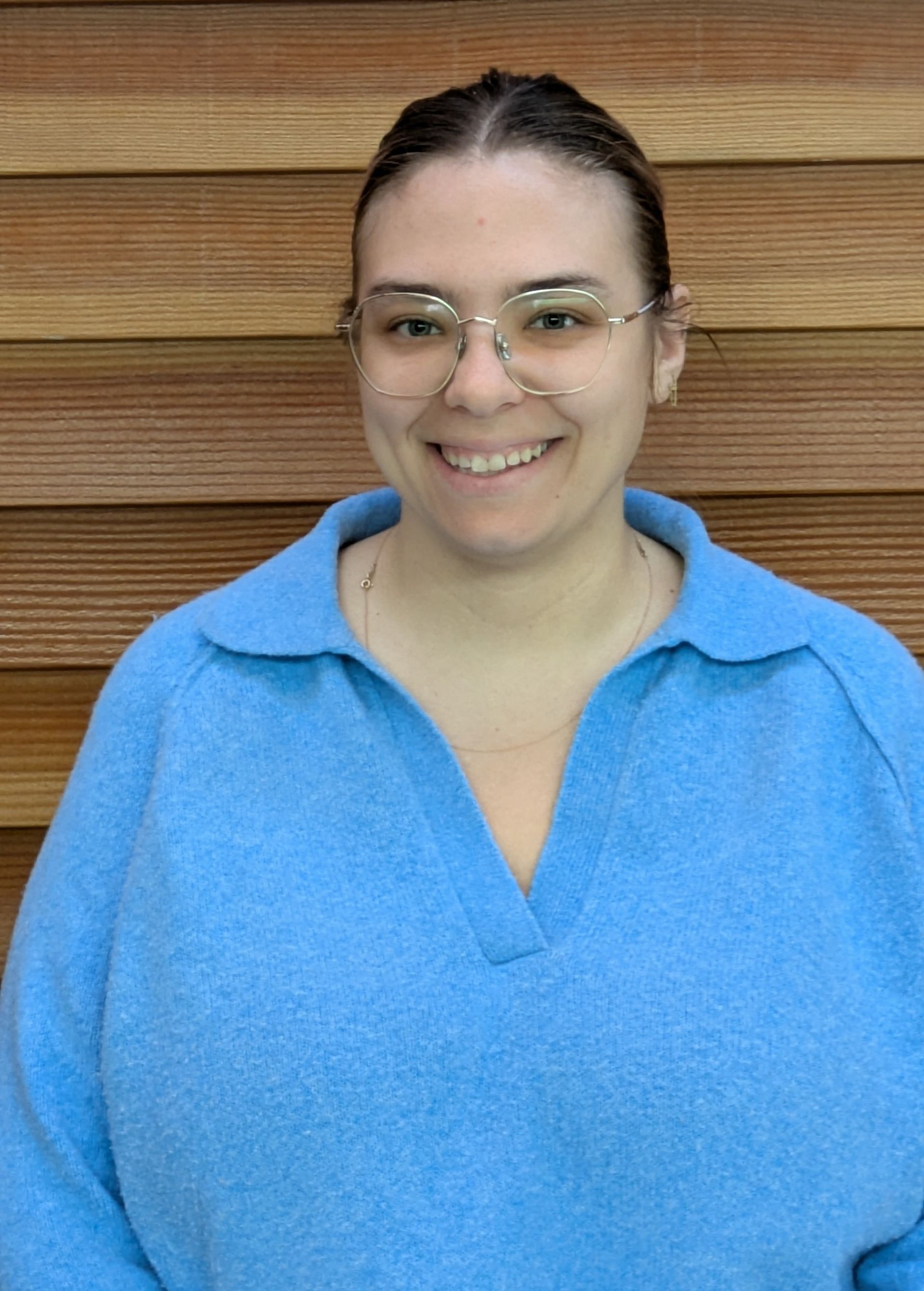 Woman with glasses smiles, wearing a light blue sweater, in front of a wood-paneled wall.