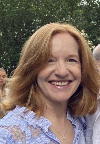 Woman with reddish-blonde hair smiles broadly, wearing a light blue top with a lace design; outdoors.