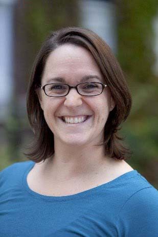 Woman with glasses smiles broadly, wearing a blue shirt, outside.