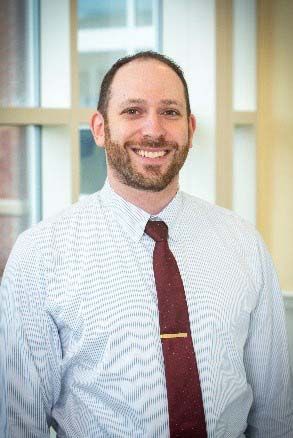Man with a beard, wearing a patterned shirt and burgundy tie, smiling in front of a window.