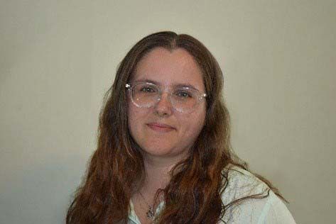 Woman with long brown hair and glasses smiling in front of a neutral-colored wall.