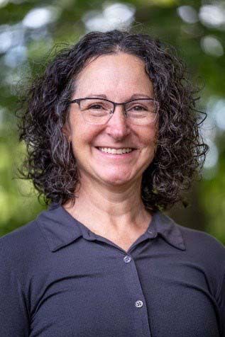 Woman with curly gray hair and glasses smiles outdoors.