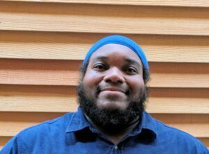 Man with a full beard and blue cap smiles at the camera, wearing a blue shirt, wood panel background.
